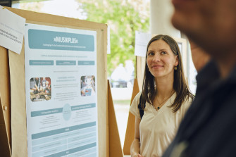 Eine junge Frau steht lächelnd vor einem Poster mit dem Titel „MusikPlus – Studiengänge für das Grundschullehramt Musik an der Musikhochschule Lübeck“. Im Hintergrund ist eine freundliche, helle Umgebung mit weiteren Postern und Personen zu erkennen.