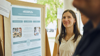 Eine junge Frau steht lächelnd vor einem Poster mit dem Titel „MusikPlus – Studiengänge für das Grundschullehramt Musik an der Musikhochschule Lübeck“. Im Hintergrund ist eine freundliche, helle Umgebung mit weiteren Postern und Personen zu erkennen.